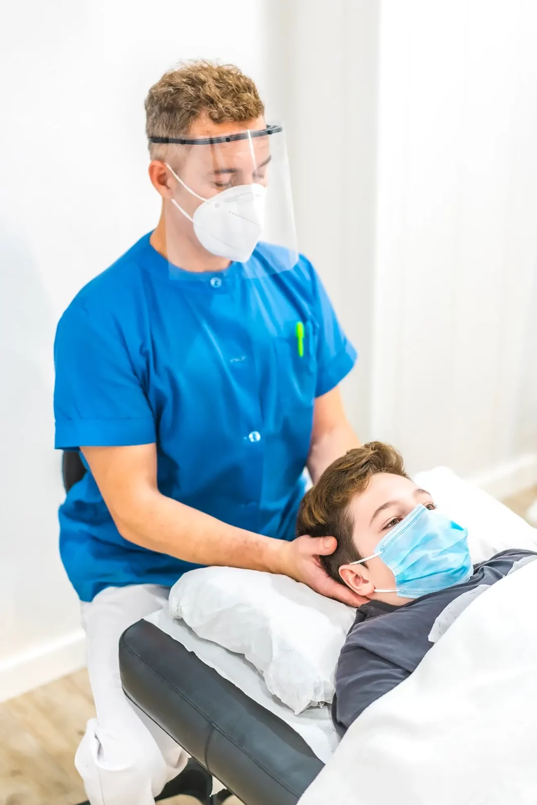 Chiropraktiker behandelt den Nacken eines Jungen; beide tragen Masken in der Physiotherapiepraxis in Heilbronn.